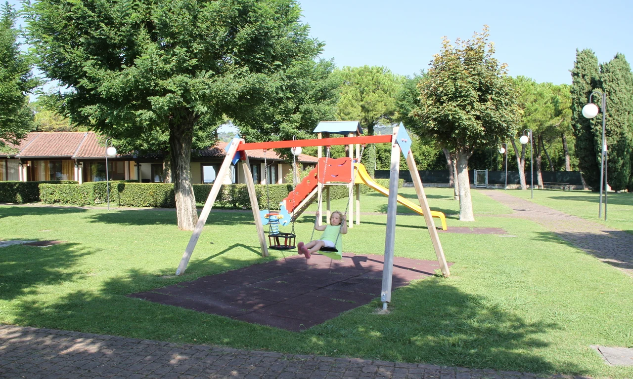 Child swinging on playground swing set; slide and climbing structure surrounded by green grass and trees at Front Lake Resort Le Corti del Lago