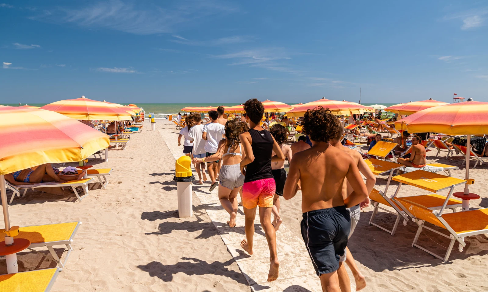 People running on a sandy beach lined with orange umbrellas and sun loungers under a clear blue sky at International Riccione Family Camping Village