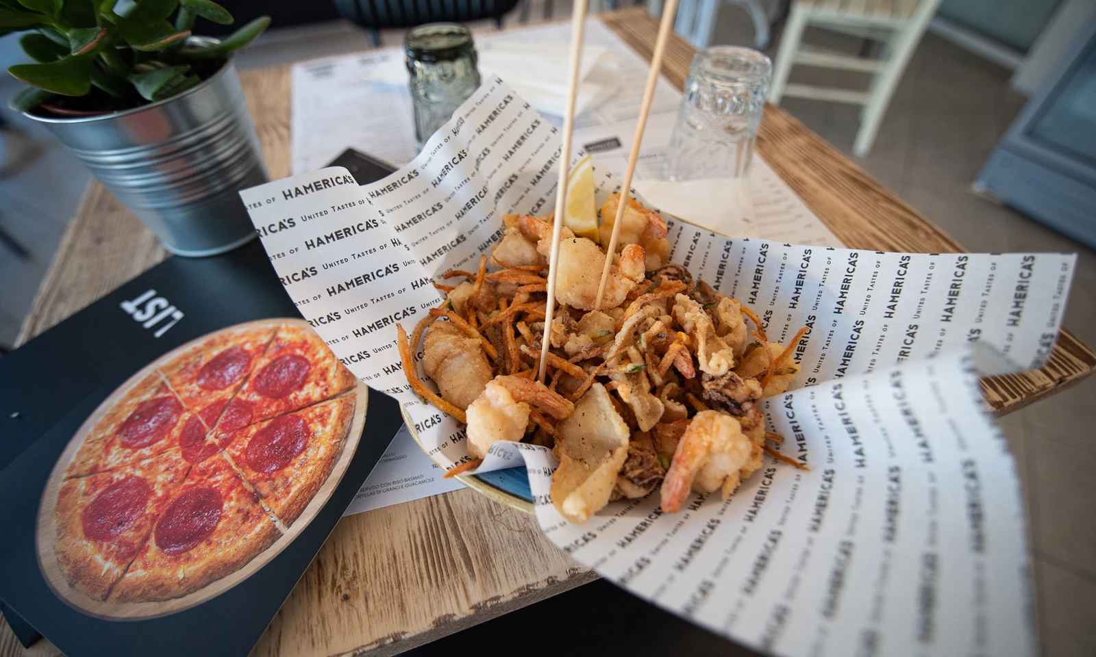 Fried mixed seafood sits on paper with skewers in a restaurant, beside a menu and plant at International Riccione Family Camping Village