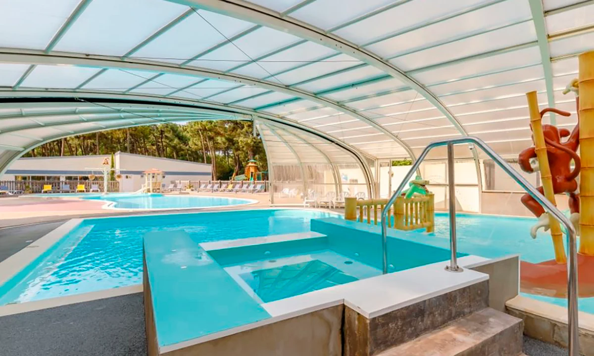 A large covered swimming pool area with steps leading into a hot tub, surrounded by lounge chairs and a playground at Camping 2 Plages & Océan Village Corsaire