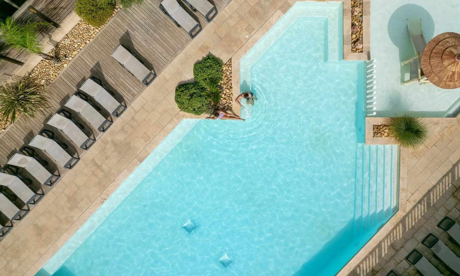 Two individuals relaxing at a swimming pool surrounded by lounge chairs and greenery at La Barque