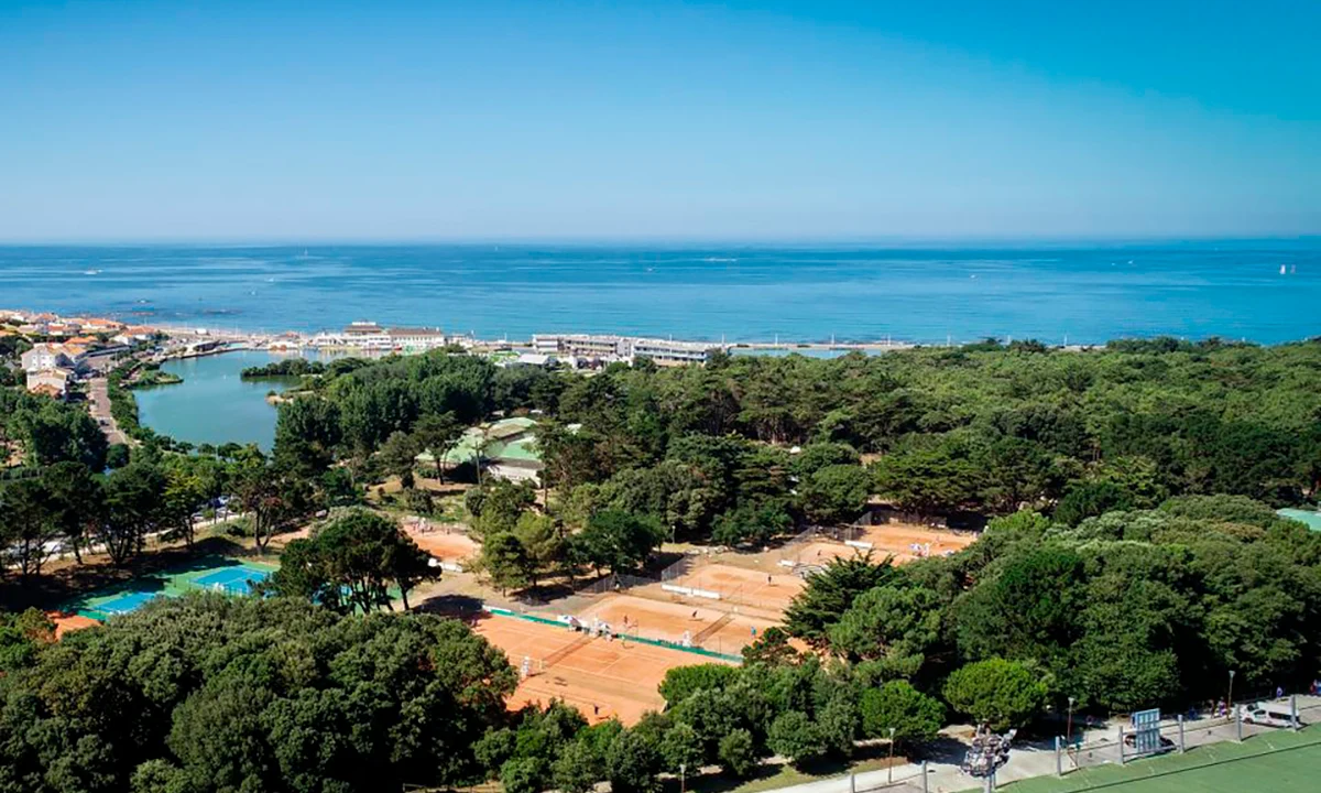 Tennis courts nestled among lush trees, situated near a small lake and buildings with the ocean in the background at Camping 2 Plages & Océan Village Corsaire