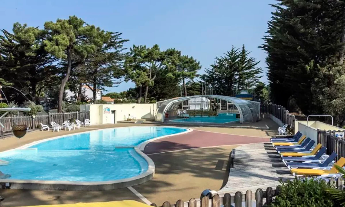 Swimming pool with surrounding lounge chairs, partially covered by structure, nestled among tall trees at Camping 2 Plages & Océan Village Corsaire