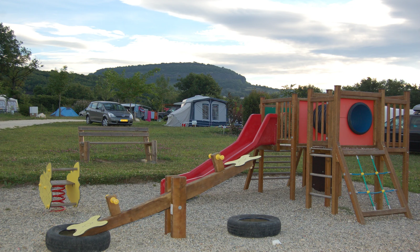 Playground equipment featuring slides, seesaw, and climbing frame, situated in a grassy campground with tents and a parked car at Les Arches