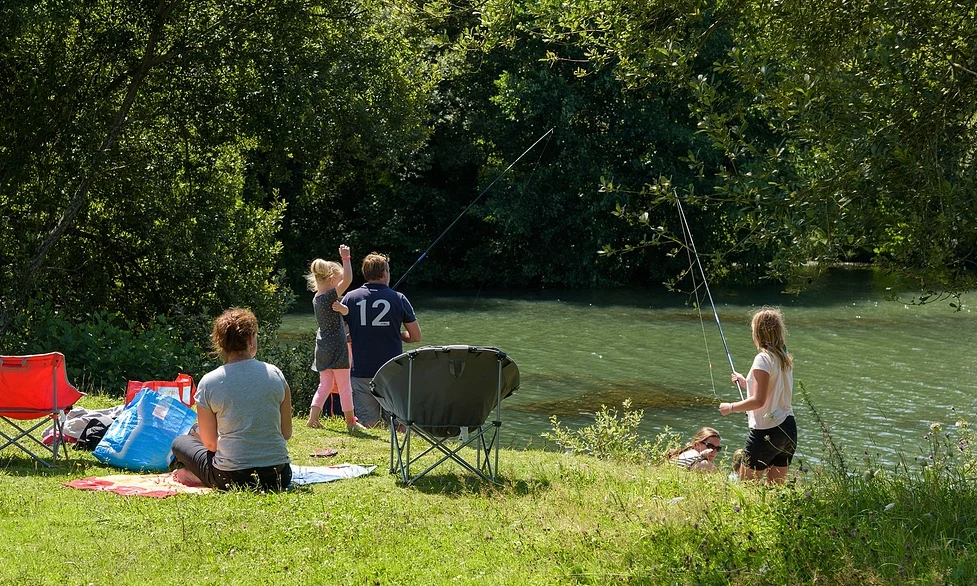 People fishing and relaxing by a riverside, surrounded by trees and grassy area with chairs and blankets at Domaine de Mesqueau