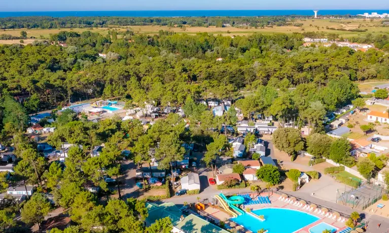 Aerial view of a campsite, featuring a large swimming pool and surrounding cabins, bordered by dense forest and a distant coastline at Le California