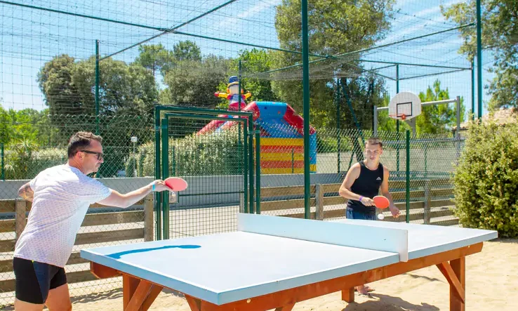 Two people playing table tennis in a fenced outdoor sports area with trees and inflatable playground in the background at Le California