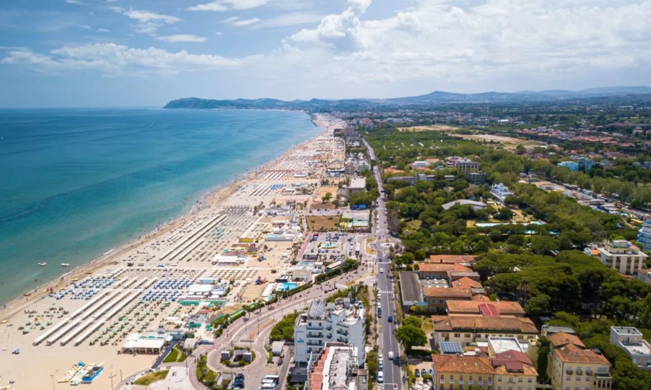 Aerial view of a long, sandy beach with organized umbrellas beside a vibrant town and lush greenery at International Riccione Family Camping Village