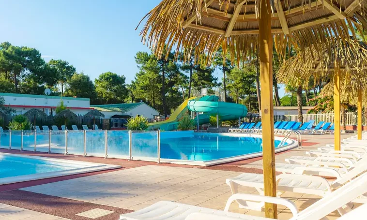 Swimming pool with water slide and lounge chairs under grass umbrellas in a forested campsite setting at Le California