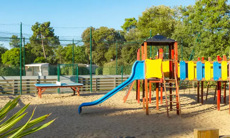 Colorful playground equipment with slide on sandy ground, surrounded by trees and sports facilities at Le California