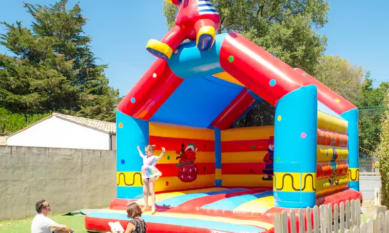 Bouncy castle with children playing, while adults watch nearby on a grassy area, surrounded by trees and buildings at Le California