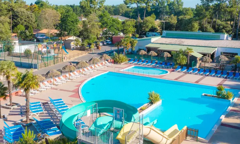 Large swimming pool surrounded by numerous sunbeds and straw umbrellas, adjacent to water slides and playground in a wooded area at Le California