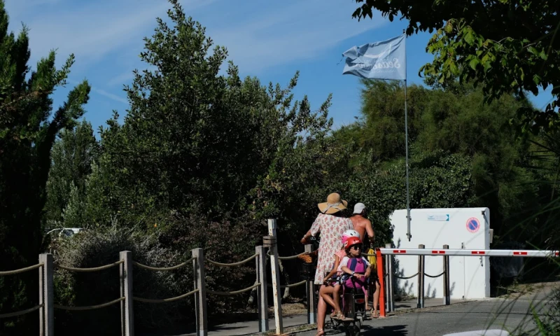 A person rides a bicycle with a child passenger, surrounded by trees and a campsite gate at Seasonova Ile de Ré