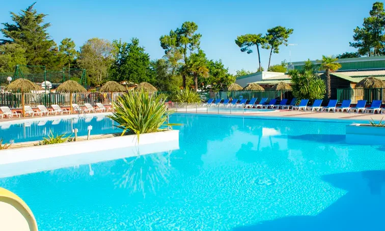 A clear blue swimming pool reflecting the sky, surrounded by sun loungers, umbrellas, and greenery at Le California