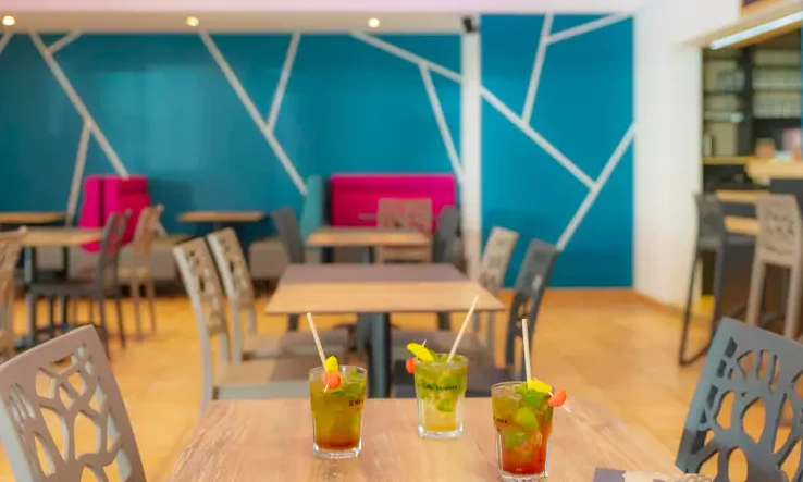 Three colorful cocktails rest on a wooden table in a modern café with geometric wall designs and vibrant seating at Le California