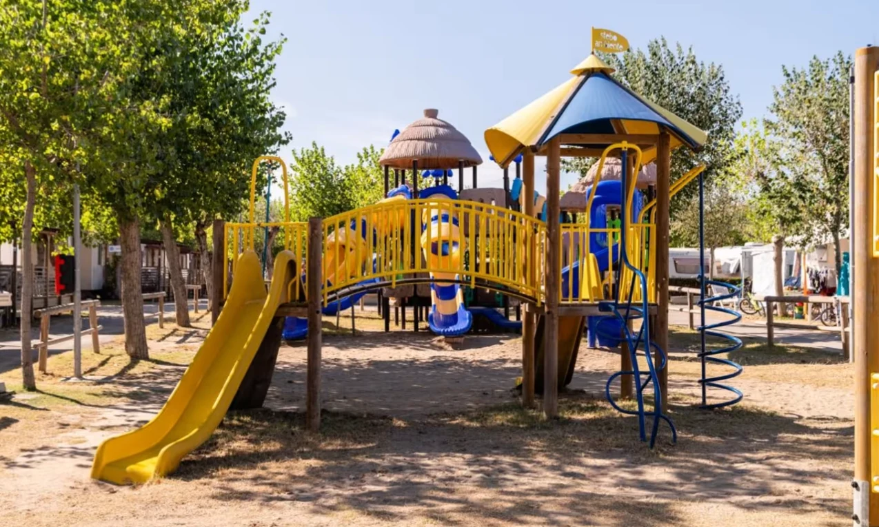 Colorful playground set stands amid trees with slides, climbing structures, and a bridge at International Riccione Family Camping Village