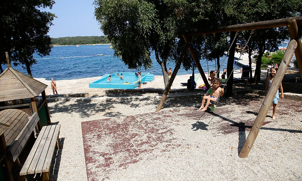 Children play on swings; background shows a beach with people swimming in a pool by the sea at Orsera