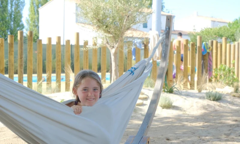 Girl relaxing in a hammock near a wooden fence, with people and buildings in the background at Seasonova Ile de Ré