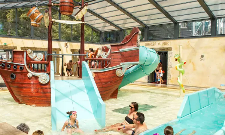 Children play on a pirate ship waterslide in an indoor pool while adults lounge in shallow water at Le California