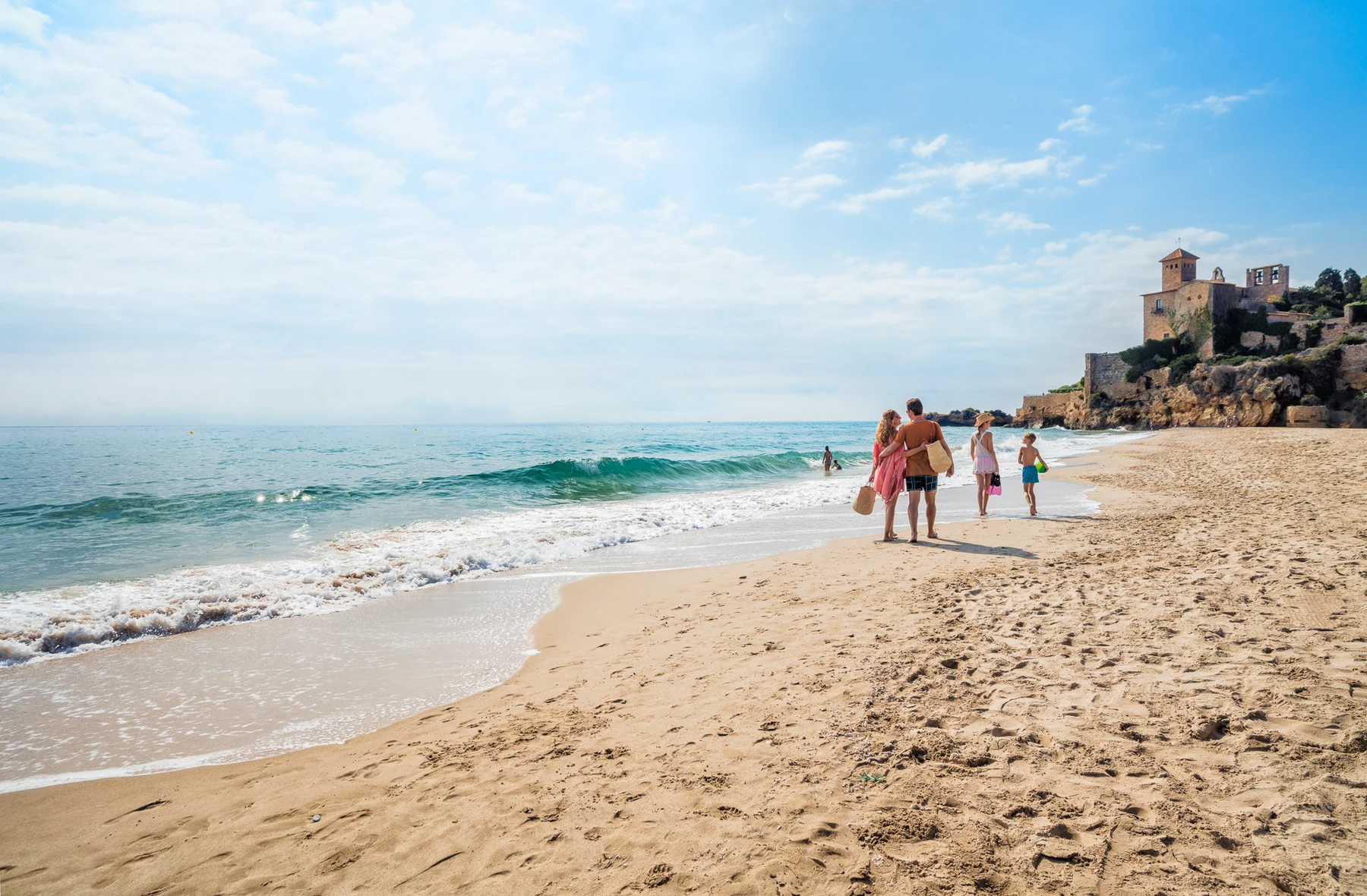People walking along a sandy beach, ocean waves beside them, with a historic building on the cliff at Tamarit Beach Resort