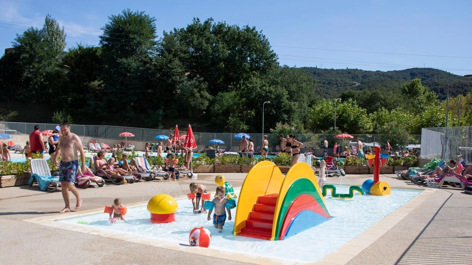 Children playing in a shallow pool with colorful play structures, surrounded by sunbathers on loungers amidst a green, hilly backdrop at La Garenne 
