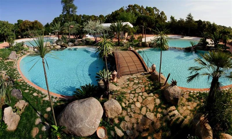 Bridge spanning a lagoon-style swimming pool with stone pathway, surrounded by lush greenery and trees at Ultima Spiaggia