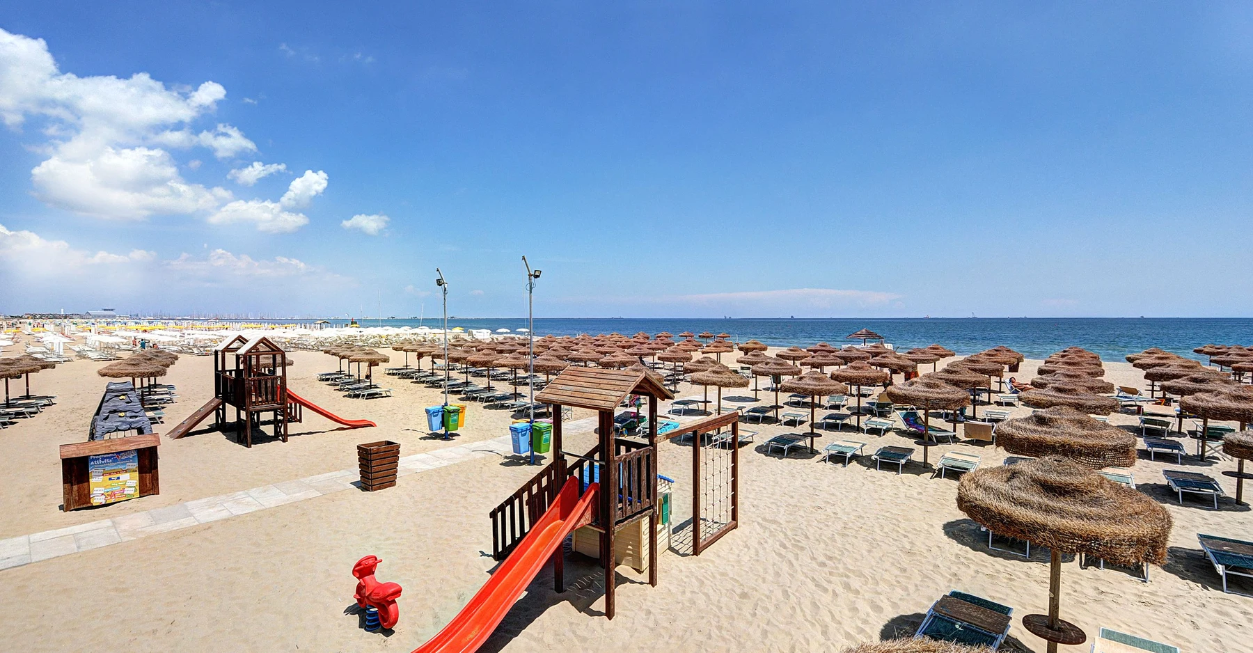 Wooden playground with slides stands on sandy beach, surrounded by straw umbrellas and deck chairs near ocean at Rivaverde Family Camping Village