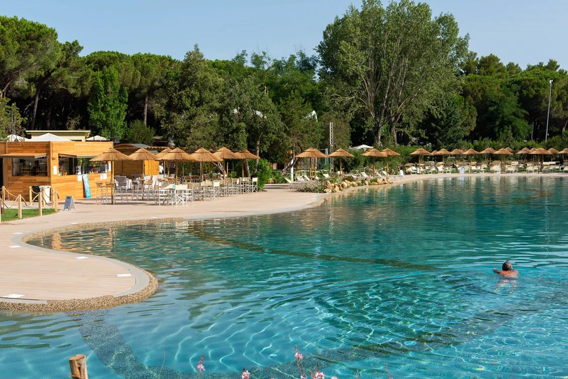 A person swims in a large, clear pool with shaded seating and dense forest in the background at Le Esperidi