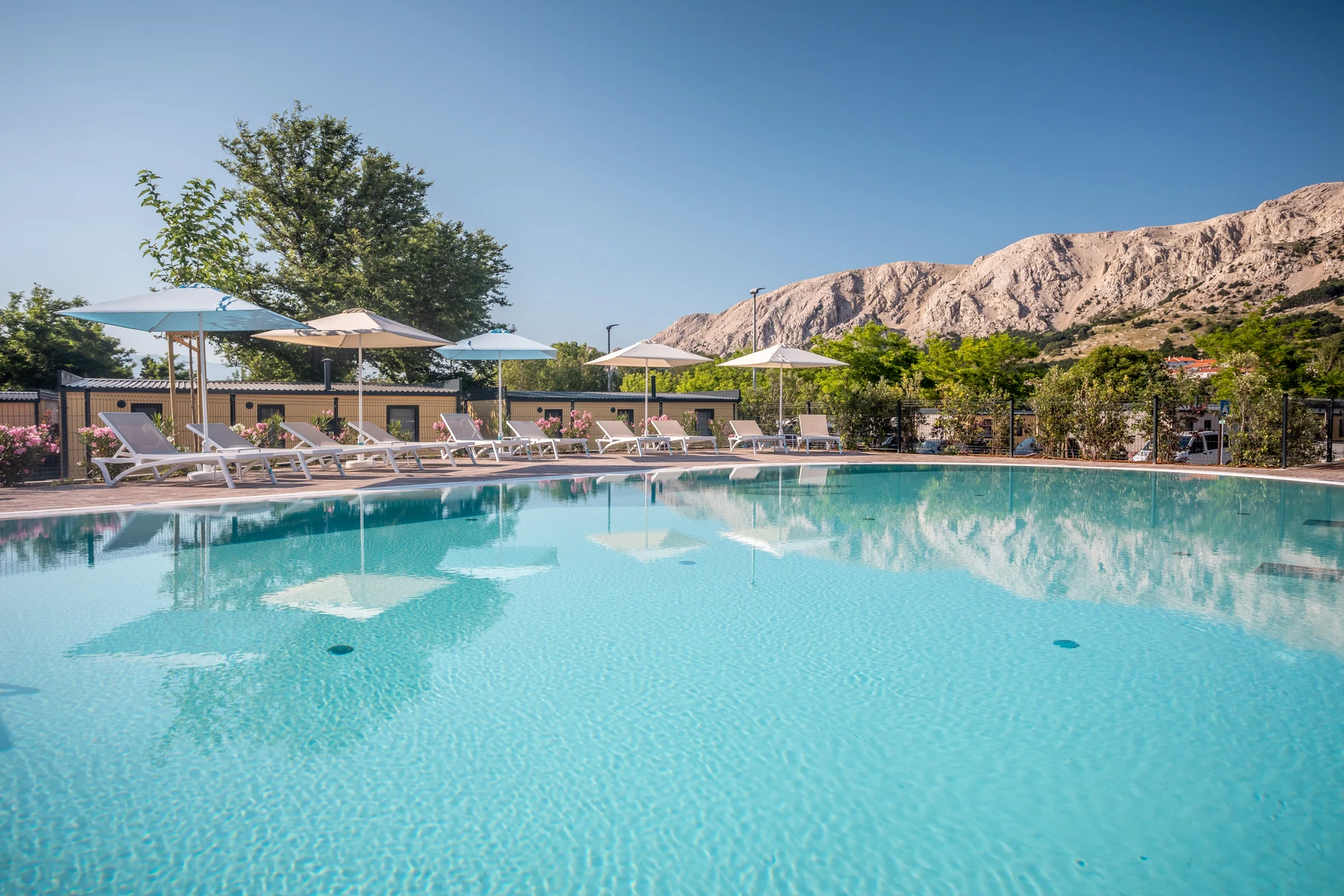 Pool with lounge chairs under umbrellas, surrounded by trees and mountains in the background at Baska Beach Camping Resort