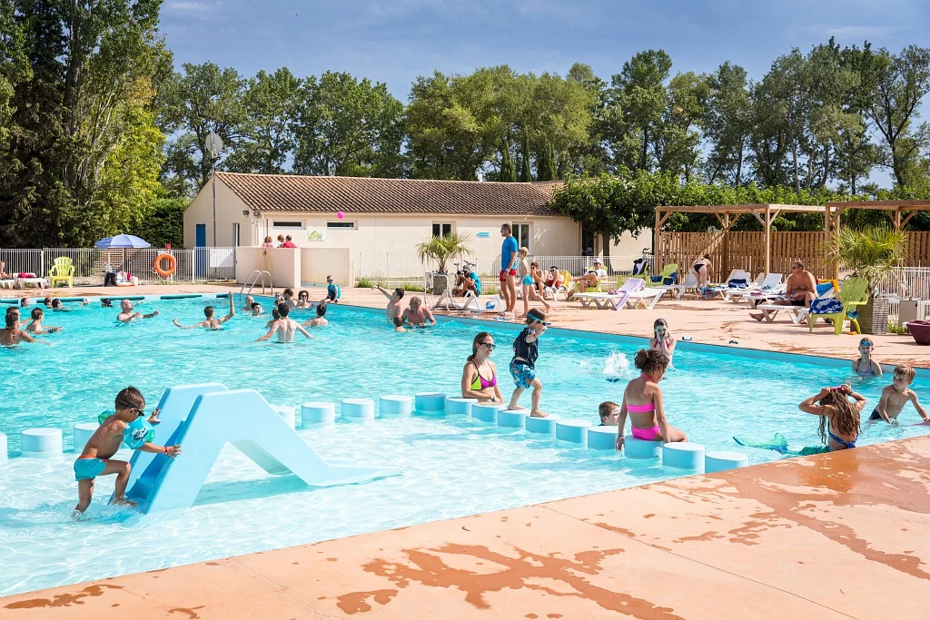 Children playing in a swimming pool with adults relaxing in lounge chairs under a clear sky at Marvilla Parks Les Rives du Luberon