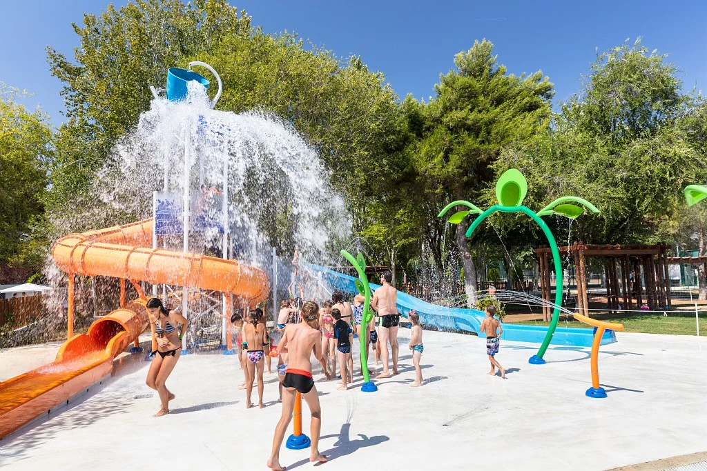 Children are playing under a large water bucket and on water slides at a colorful splash pad, surrounded by trees at Tamarit Beach Resort