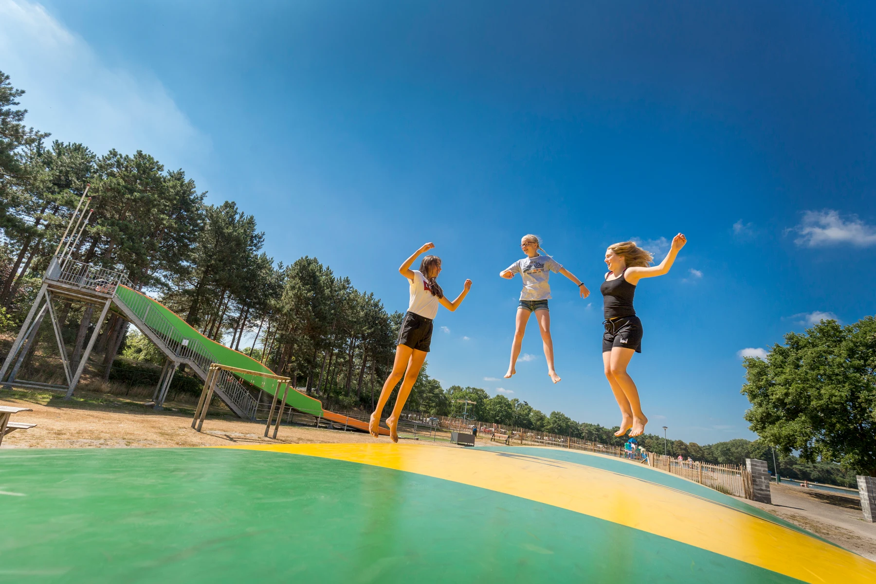 Three people are jumping on a colorful inflatable cushion near slides and trees at de Bergen