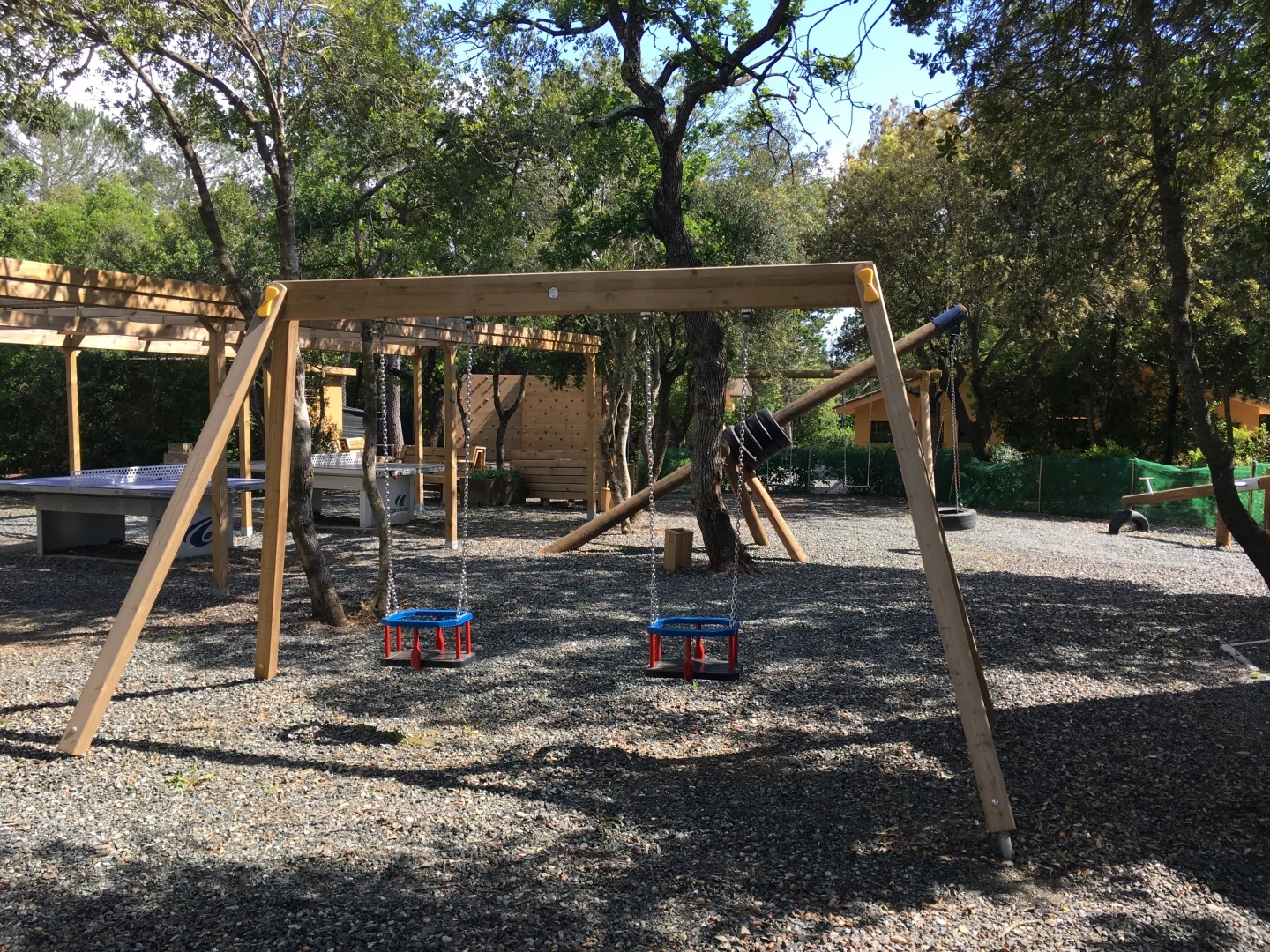Wooden swing set with blue seats under trees, surrounded by play structures and activity tables at hu Montescudaio Village