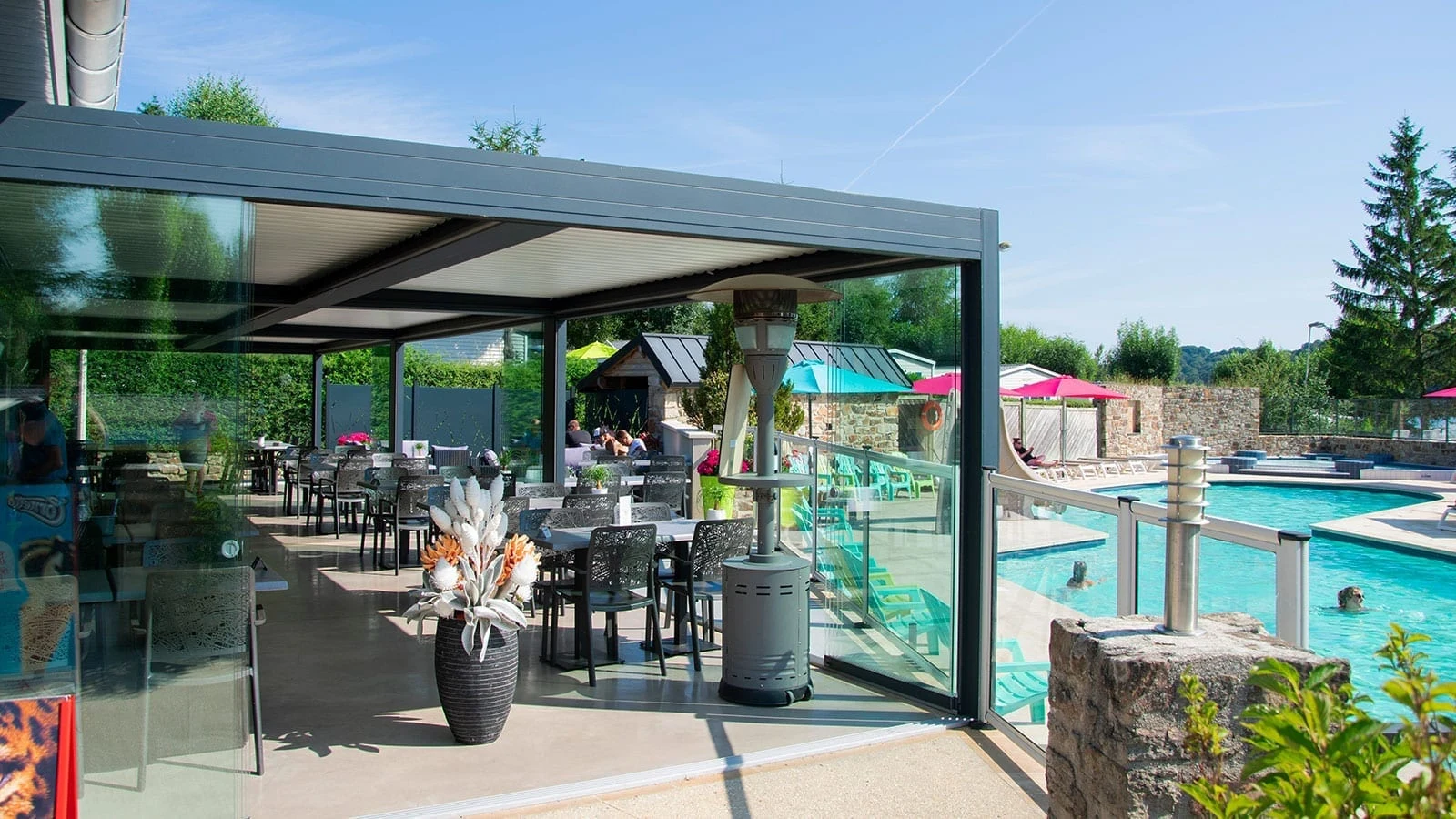 Patio dining area with glass walls, overlooking a swimming pool with people swimming, greenery and blue sky at Les Genêts 