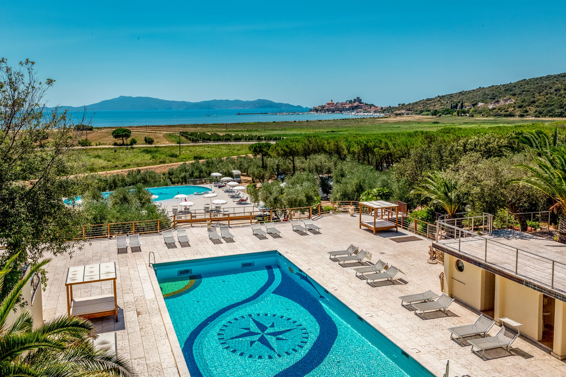 Swimming pool surrounded by loungers; distant coastal town, lush greenery bordering landscape. Sunny outdoor setting at Gitavillage Talamone