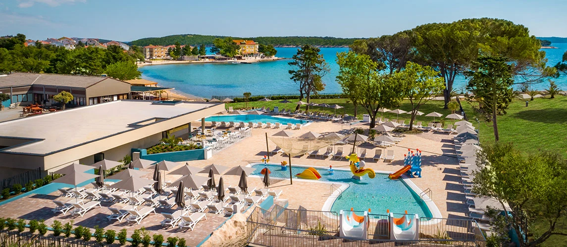Pool with slides surrounded by sun loungers, trees, and umbrellas near a scenic lake at Valamar Camping Padova
