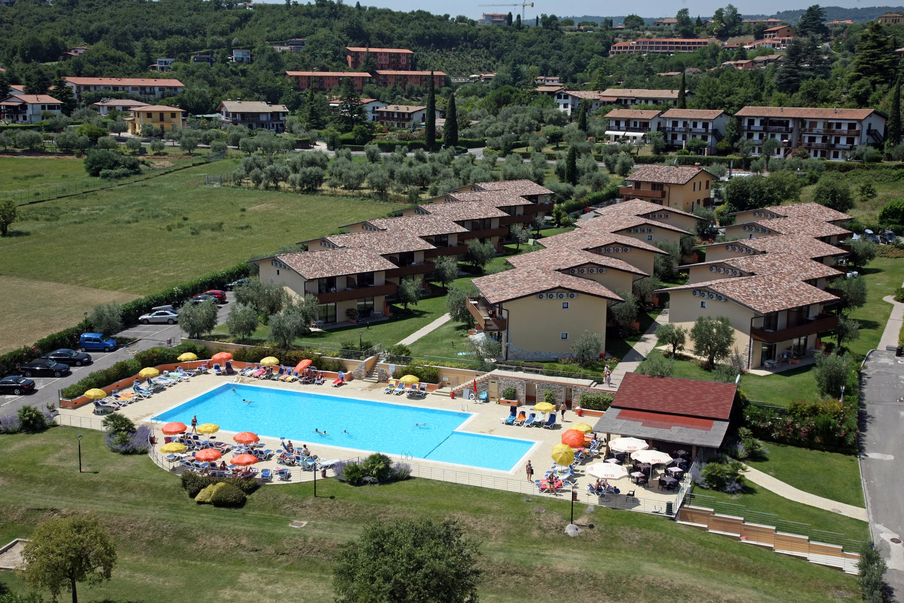 Swimming pool surrounded by umbrellas and sunbathers, adjacent to multiple buildings, nestled in countryside with hills and houses at Onda Blu Resort