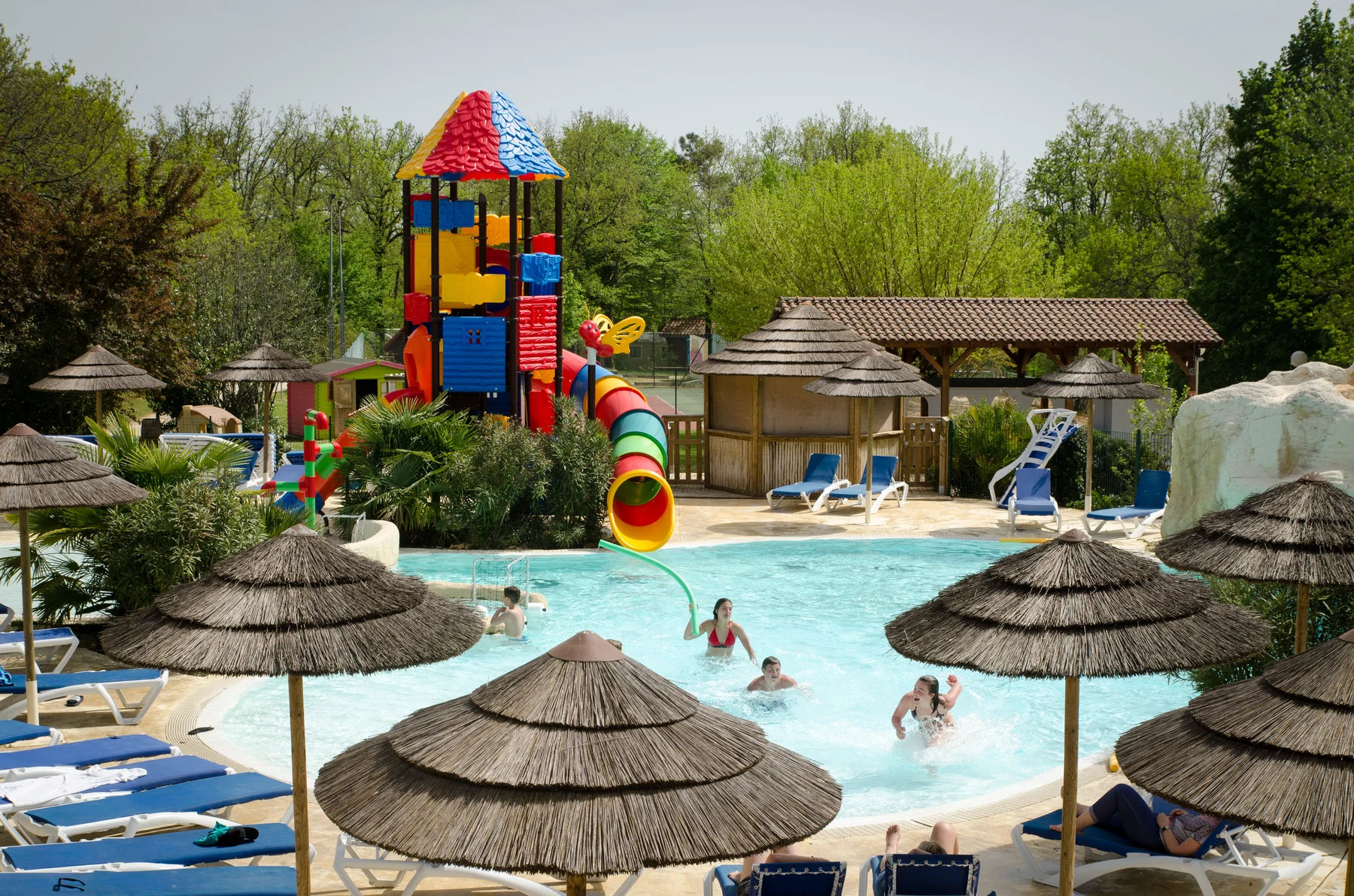 Children play in pool near colorful water slide tower, surrounded by lounge chairs and thatched umbrellas at l'Évasion