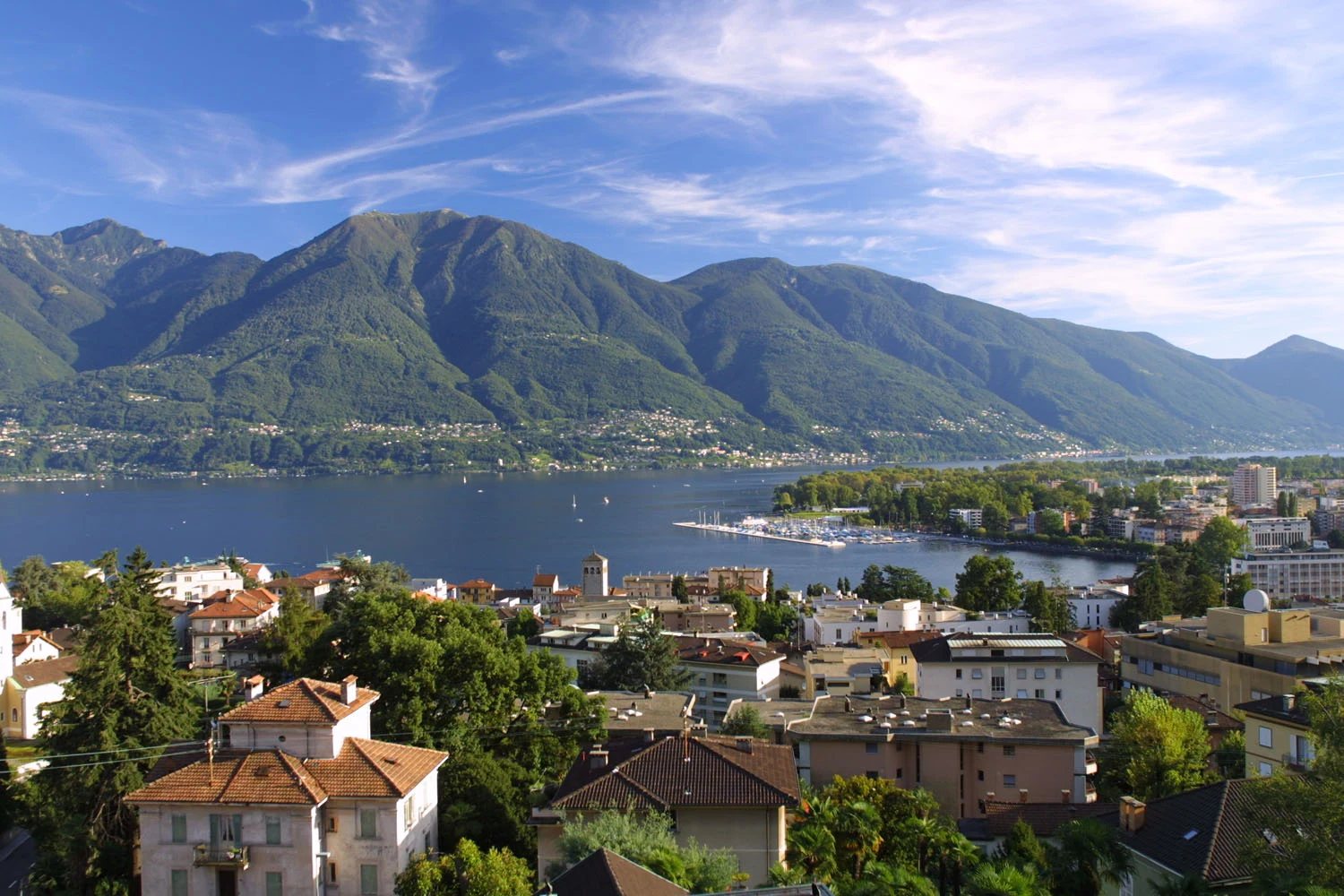 Buildings overlook a serene lake with surrounding lush green mountains under a partly cloudy blue sky at Lido di Monvalle