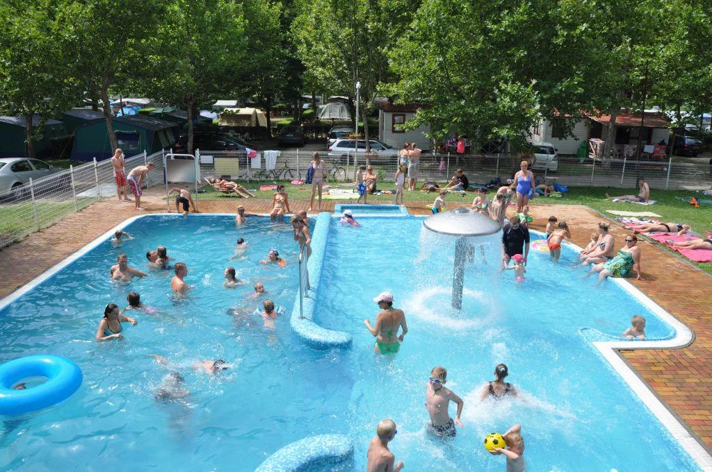 People swimming and playing in a crowded pool with a water feature, surrounded by trees and camping tents at Pelso