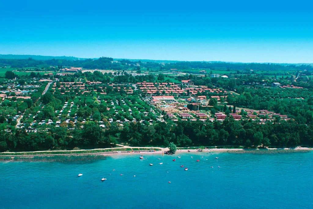 Aerial view of a large campsite near a sandy beach with boats in the water, surrounded by green landscape at Piani di Clodia