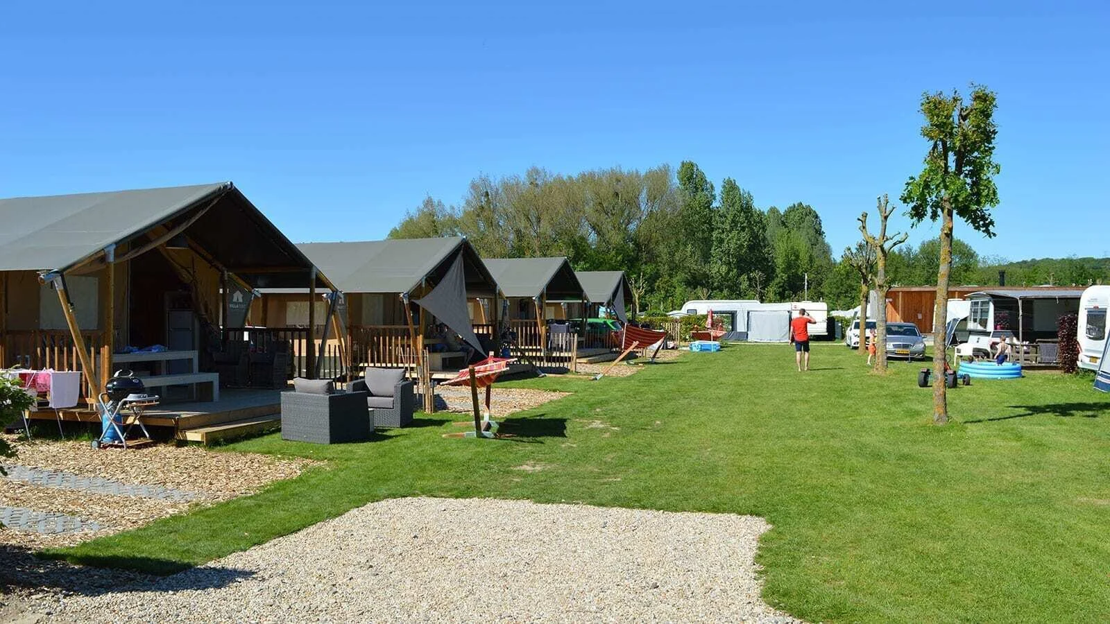 Cabins and tents line grassy campsite with scattered chairs, hammock, and a man walking; cars and trailers in background at 't Geuldal
