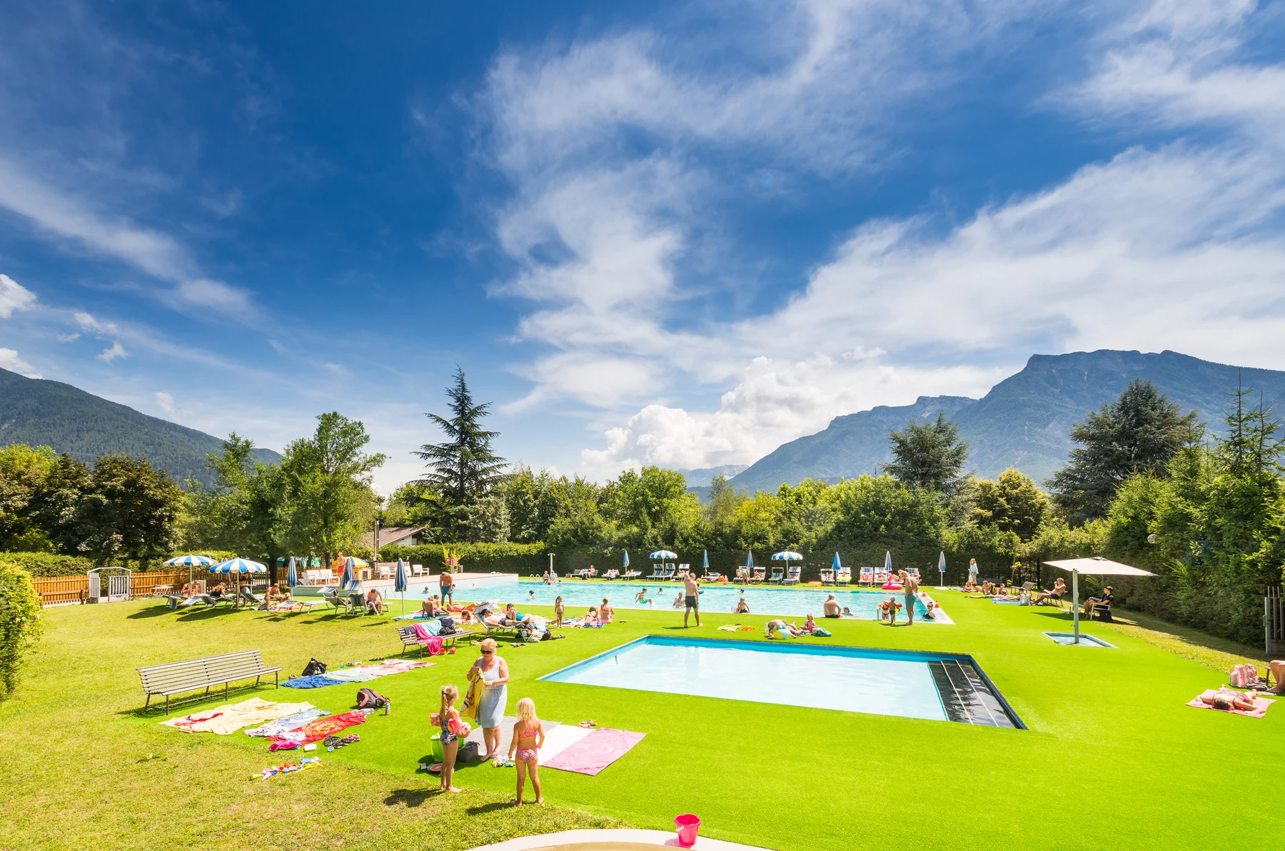 People sunbathing and swimming poolside in a grassy area, surrounded by trees and mountains under a vibrant blue sky at Due Laghi