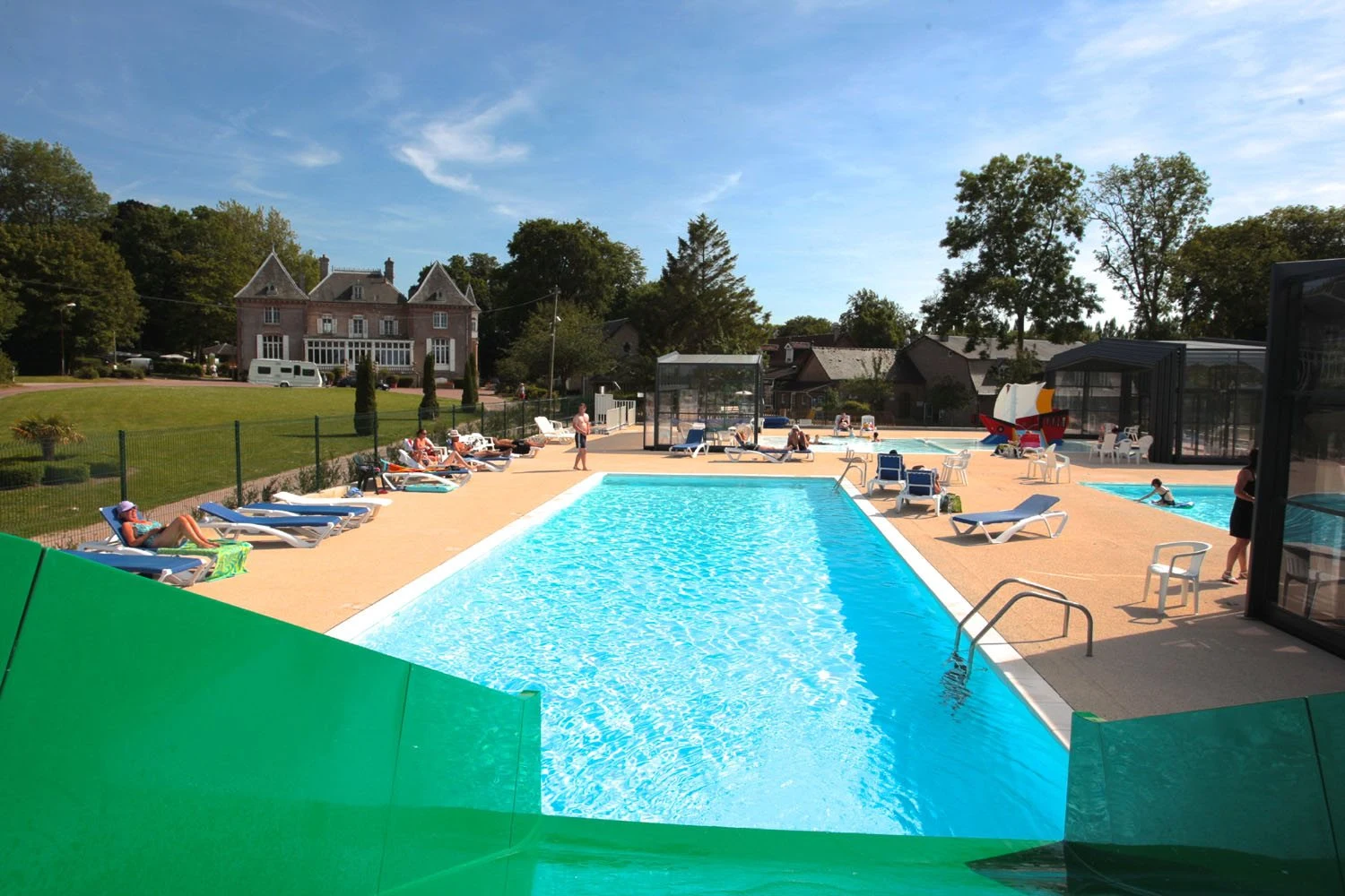 People relax on loungers beside a large outdoor swimming pool in a scenic area with historic buildings and greenery at Domaine du Chateau de Drancourt