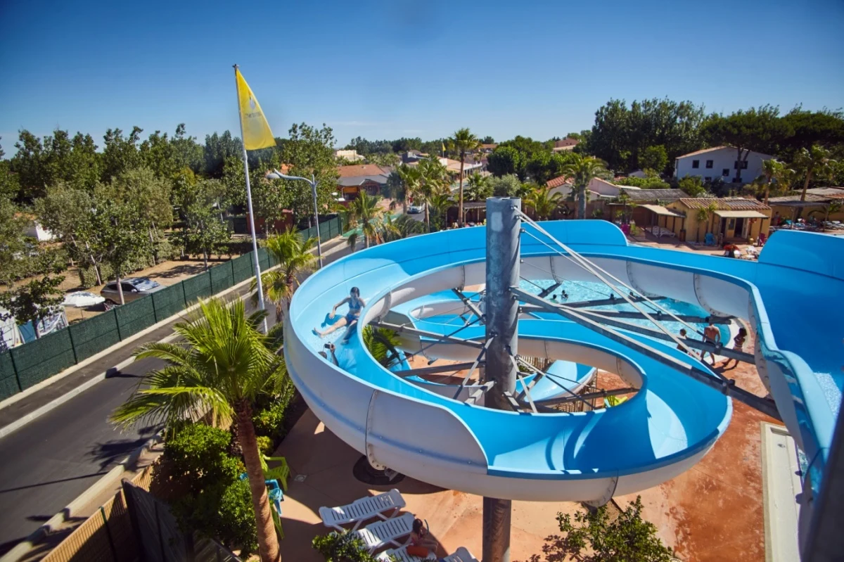Person sliding down a blue water slide, surrounded by palm trees and buildings at Les Mediterranées Nouvelle Floride