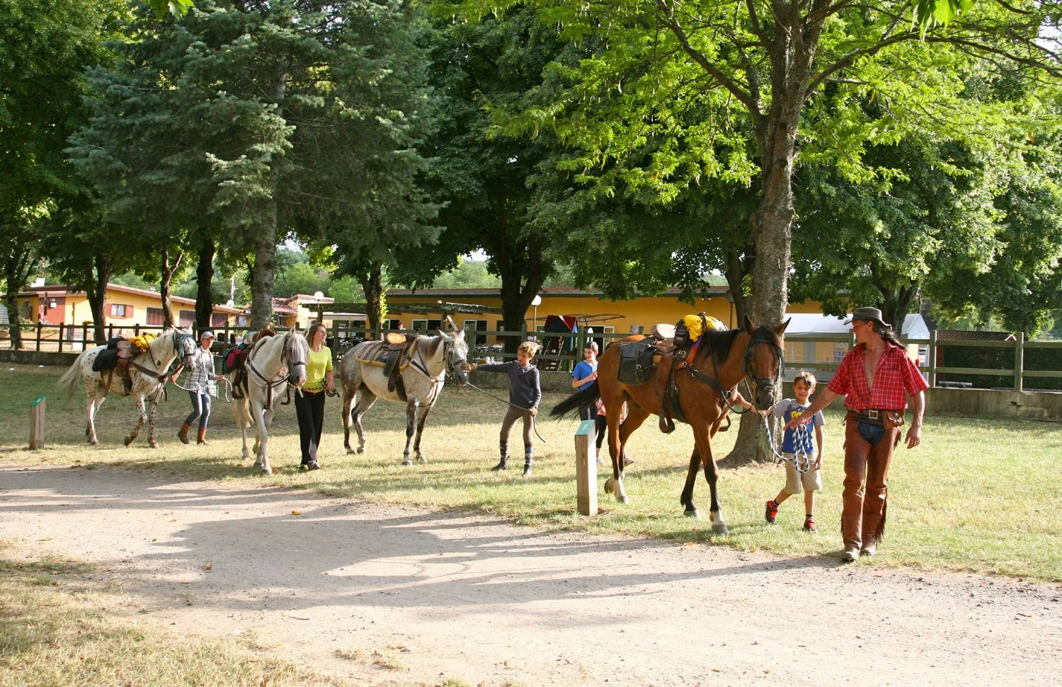 People leading horses in a tree-filled camping area with buildings in the background at Le Ranch des Volcans