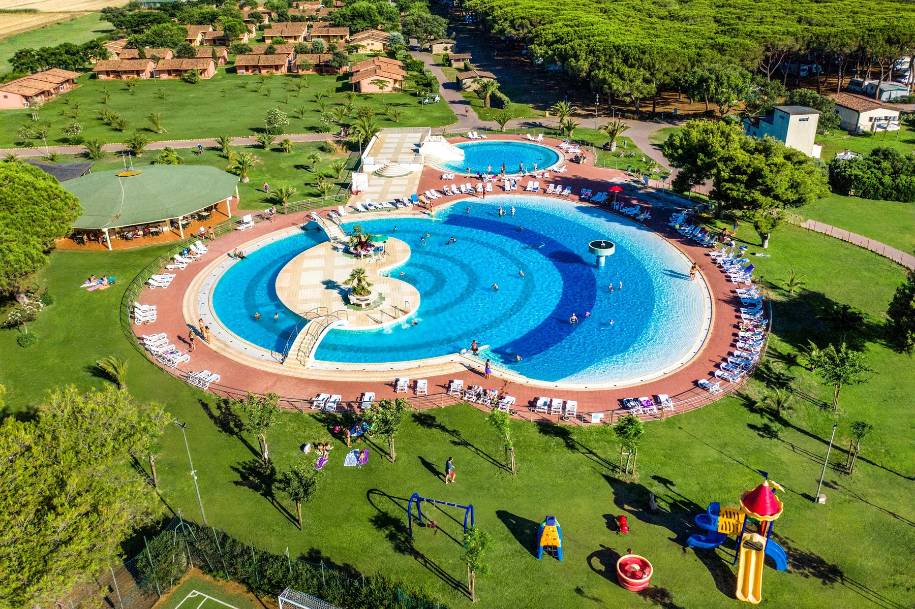 Large outdoor pool with people swimming, surrounded by lounge chairs and grassy areas; playground and buildings nearby at Gitavillage California