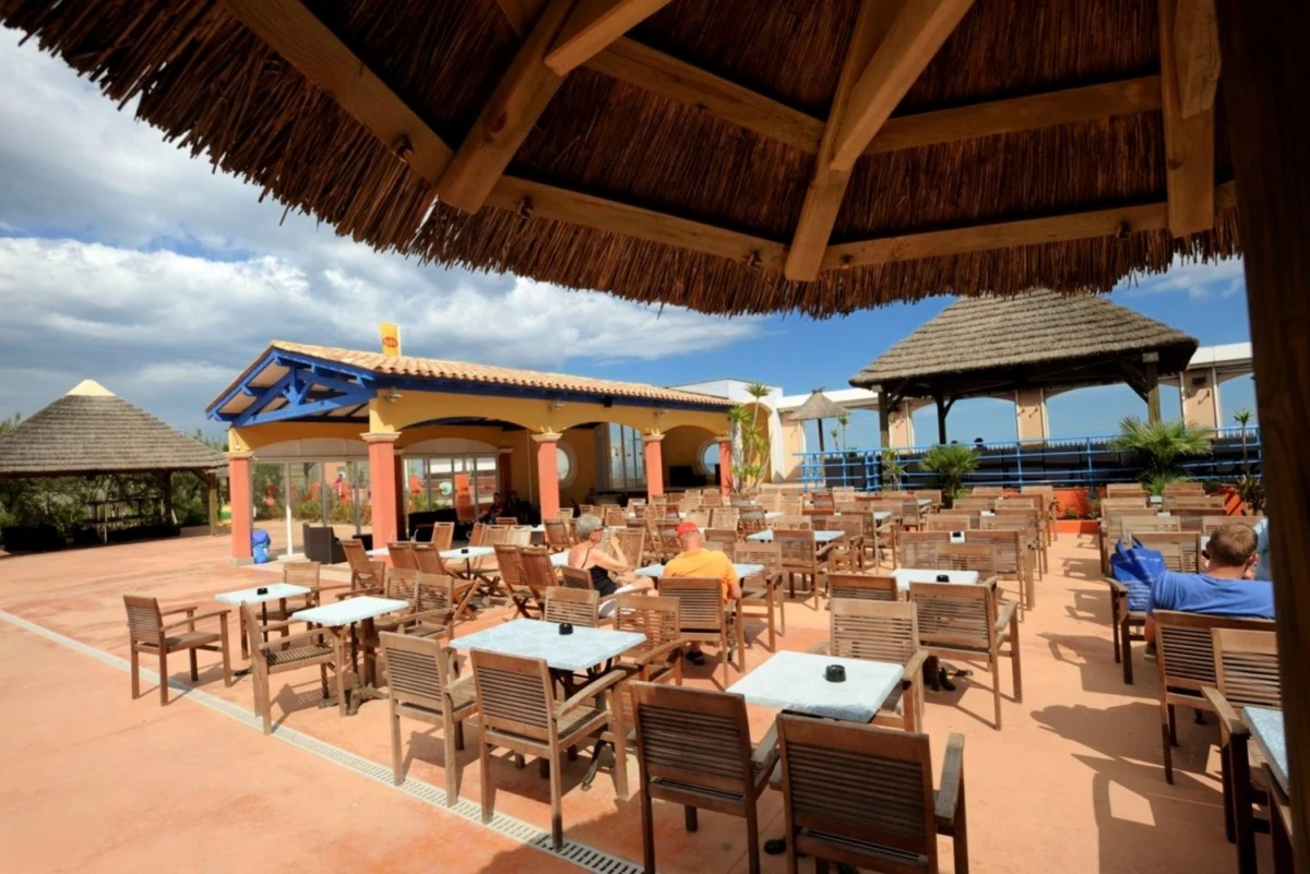 People sitting at wooden tables under thatched roofs, surrounded by colorful buildings and a sunny, open-air setting at Les Mediterranées Nouvelle Floride