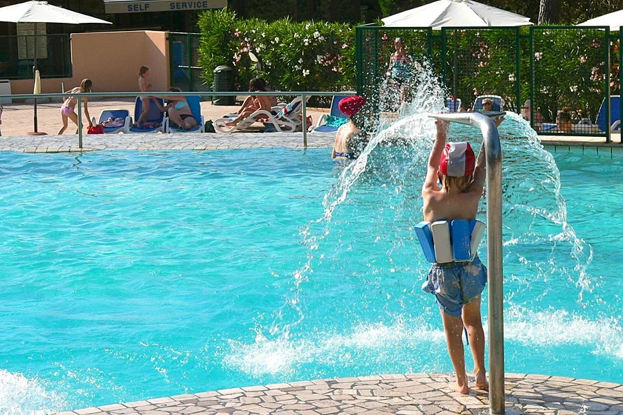 Child plays with pool fountain, wearing a flotation device, surrounded by other children and adults lounging poolside at Ville degli Ulivi