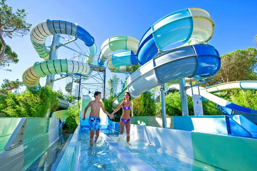 Two people walk hand in hand beneath a twisting water slide surrounded by greenery at Marvilla Parks Le Castellas